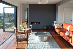 Antique chair and leather sofa around round coffee table on rug in front of fireplace in dark grey partition wall; open folding terrace doors with view of wooded landscape