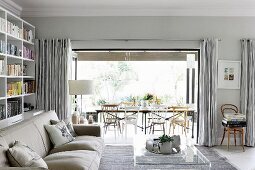 Pale sofa and transparent coffee table in modern living room with rustic ambiance; open folding doors with view of long table and chairs in background