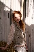 A young woman wearing a lace blouse and a leather gillet standing in front of a corrugated iron wall