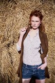 A young woman wearing denim shorts, a floral-patterned blouse and a leather gillet standing by bales of hay