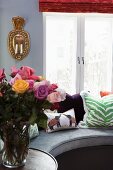Vase of roses on side table next to curved bench with arranged scatter cushions in front of window