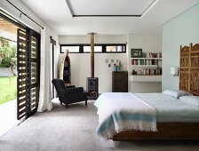 Double bed with carved wooden panel on wall and armchair in front of log-burner in bedroom with open terrace doors