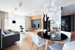 Dining table with black, round top and white chairs below Zettel'z lamp in open-plan interior with lounge area