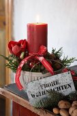 Brennende Kerze mit weihnachtlichem Gesteck in einer Keramikschale, Zinkschild und Geschenkband mit Weihnachtsgruß
