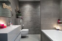 Elegant bathroom with grey wall tiles of various formats and lit candles on washstand with trough-style sink