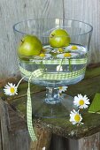 Green apples and ox-eye daisies in bowl of water decorated with green gingham ribbon on vintage wooden bench