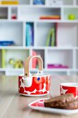 Small teapot, cup and plate of cake on dining table; bookcase in background