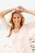 A blonde woman wearing a pastel pink three-quarter sleeved shirt on a beach holding her arms above her head