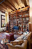 Sofa and small, upholstered footstool used as side table on rug; floor-to-ceiling wooden bookshelves against wall in background