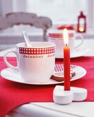 Rote, brennende Kerze im herzförmigen Kerzenhalter und Tassen mit Karo-Dekor