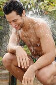 Mann in Badeshorts hockt unter Dusche im Freien