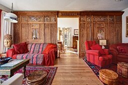 Kunsthandwerklicher Raumteiler in offenem elegantem Wohnraum; Sofa und Sessel in Rottönen, orientalische Lederpoufs mit Tisch und Ethnokunst - in einem Fertighaus