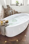 An oval bathtub filled with bubbles in a decorative, natural stone-clad corner with wooden-style floor tiles and spotlights