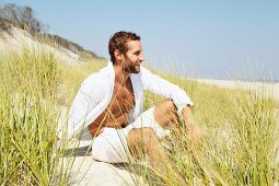 Junger Mann in weisser, luftiger Kleidung sitzt in Sanddüne am Strand