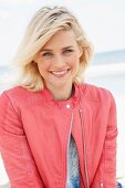 Blonde Frau mit blauem Pulli und lachsfarbener Lederjacke im Bikerstil am Meer