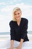 A blonde woman on a beach wearing a dark woollen jumper and white trousers