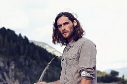Junger Mann mit Bart und langen Haaren vor bewaldetem Berg