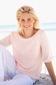 A smiling blonde woman on a beach wearing a pastel-coloured top and trousers