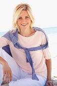 A smiling blonde woman on a beach wearing a pastel-coloured top, jumper and trousers