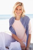 A smiling blonde woman on a beach wearing a pastel-coloured top, jumper and trousers