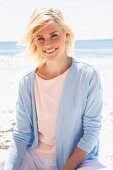 A smiling blonde woman on a beach wearing a pastel-coloured top, jacket and trousers