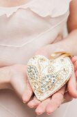 White, heart-shaped bauble with gilt ornamentation and rhinestones hald in woman's hands