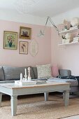 Hellgrau lackierter Couchtisch und Eckpolstersofa vor rosa getönten Wänden, oberhalb gerahmte Bilder
