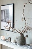 Baubles hung from contorted hazel branch and arrangement of candles on patterned tiles against grey wall