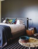 Colourful scatter cushion on double bed against dark wall; bowl on bistro table in foreground