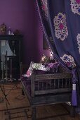 Draped curtain and antique bench with patterned scatter cushions in corner of room painted aubergine