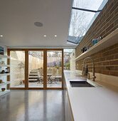 White worksurface in designer kitchen with exposed brickwork and view of terrace, steps and garden wall through terrace windows