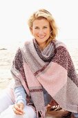 A blonde woman wearing a woollen shawl on a beach