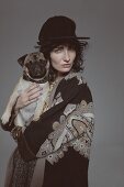 Dunkelhaarige Frau im Mantel und Hut mit Hund auf den Arm