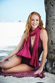 A young woman on a beach wearing a dark red bathing suit and a scarf