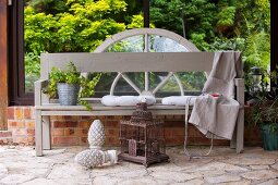 Garden bench painted pale grey on terrace against glazed side wall behind vintage birdcage and garden ornament on stone floor