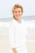 A blonde woman on a beach wearing a blue top and a white round-neck jumper