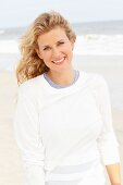 A blonde woman on a beach wearing a blue top and a white round-neck jumper