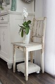White orchid on wooden chair with white peeling paint above small upholstered footstool
