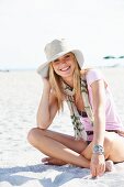 Junge blonde Frau mit Sommerhut, Schal und rosa T-Shirt sitzt im Sand am Strand