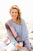 A blonde woman on a beach wearing a blue jumper and a pink-toned shawl over her shoulders