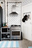 Stainless steel gas cooker below extractor hood against white-tiled wall and blue and white stripes rugs on rustic wooden floor