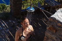 Frau in Bikini unter Dusche im Freien an rustikalem Steinhaus