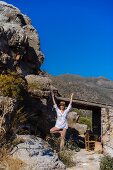 A woman practising yoga by a rustic stone house above a bay