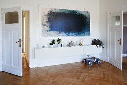 Blue painting above low sideboard against white wall in living room with herringbone parquet floor