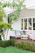 View from the garden onto a veranda with light rattan furniture in front of a wood-clad, white country house