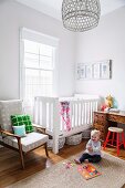 Kleinkind auf Sisalteppich vor weißem Gitterbett und Retrosessel neben Fenster in Zimmerecke