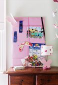 Pink pin board with drawing, calendar and boomerang behind table lamp on wooden chest of drawers