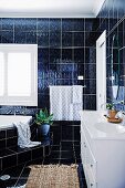 Badezimmer mit dunklen Wand- und Bodenfliesen, Eckbadewanne mit Stufe und weißem Waschtischmöbel