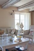 Pale sofa and antique armchair around simple white coffee table in rustic living room with whitewashed wood-beamed ceiling