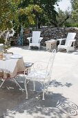 White metal chairs and breakfast table on sunny terrace with outdoor armchairs against stone wall
